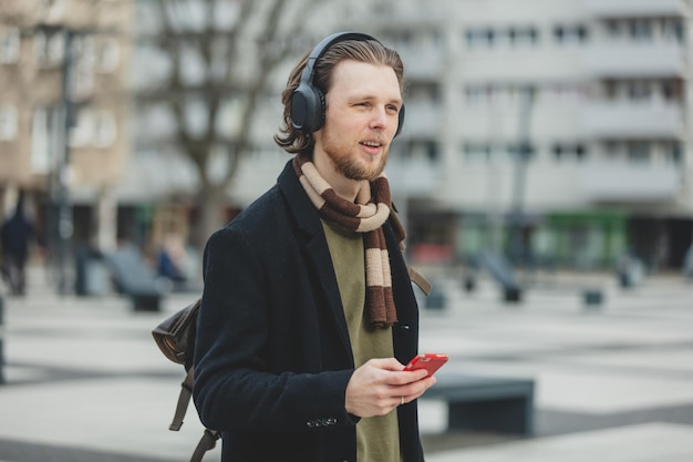 スカーフとコートを着たスタイリッシュな男が、ポーランドのヴロツワフの通りで携帯電話を使用してストリーミングサービスで音楽を聴く