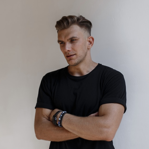 Stylish handsome American man with hairstyle in fashionable black t-shirt posing near a white wall