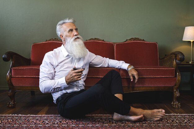 Stylish and handsome bearded senior man sitting on the floor and drinking red wine