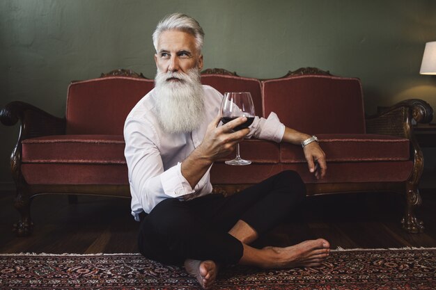 Stylish and handsome bearded senior man sitting on the floor and drinking red wine