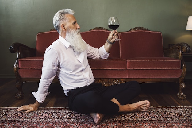 Стильный и красивый бородатый старший мужчина сидит на полу и пьет красное вино