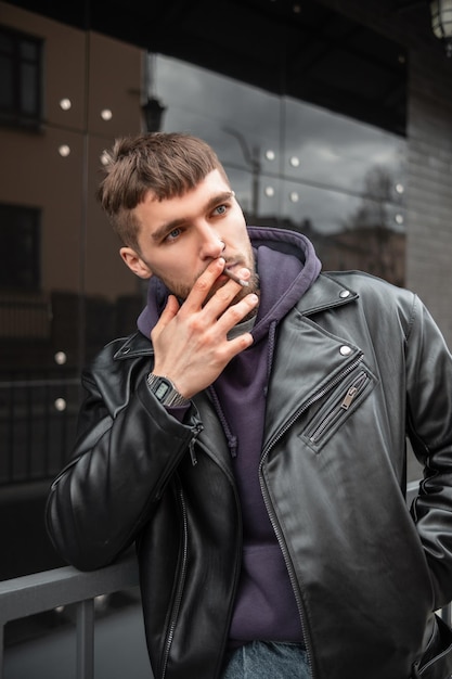 Stylish handsome brutal guy in fashionable clothes smoking a cigarette in the city