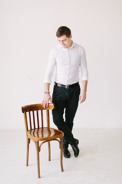 Stylish handsome groom standing in white room posing with the wooden chair