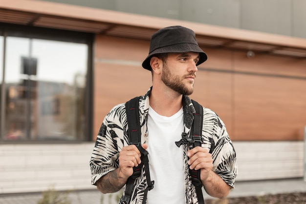 Stylish handsome hipster man with a bucket hat in fashionable clothes and a shirt with a backpack walks in the city