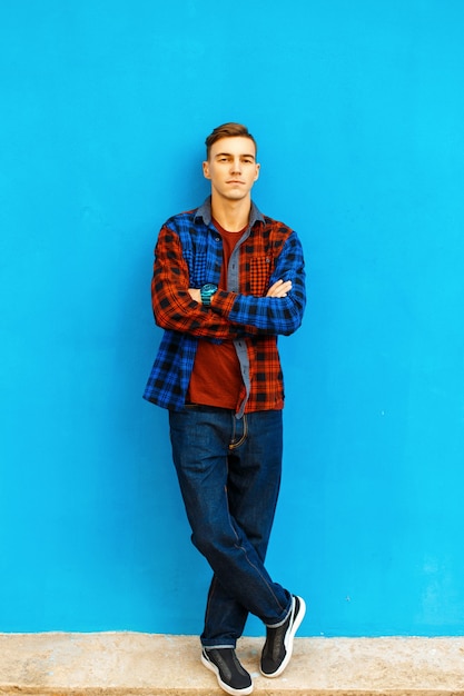 Stylish handsome man in a fashionable shirt, sneakers and jeans stands near a bright blue wall.