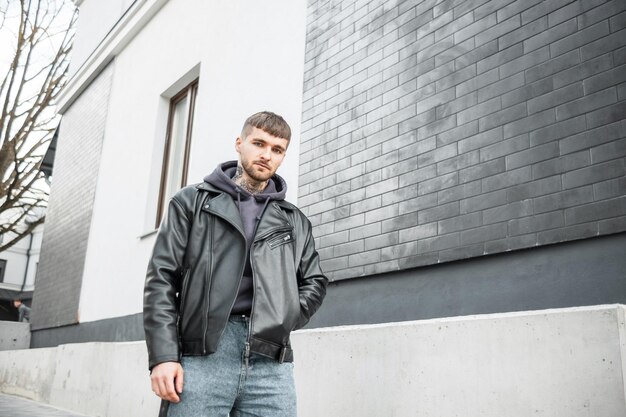 Stylish handsome man hipster in fashionable clothes walking on the street near a black brick building