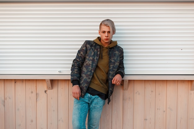 Stylish handsome man in a military jacket and jeans standing near the wall