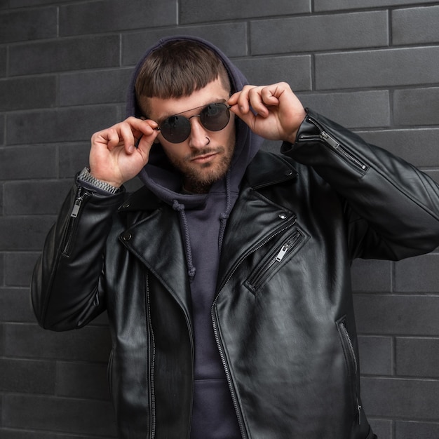 Stylish handsome man model in fashionable casual street outfit with hoodies and leather jacket wears round sunglasses and stands near a brick black wall