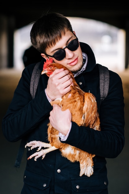 Stylish handsome young boy in sunglasses outdoor portrait. Man in black holding domestic chicken in city street.