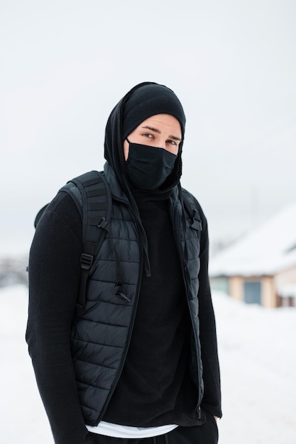 Elegante bel ragazzo in abiti neri alla moda con un giubbotto, un cappello e uno zaino viaggia e cammina all'aperto con la neve. coronavirus e concetto di stile invernale maschile urbano