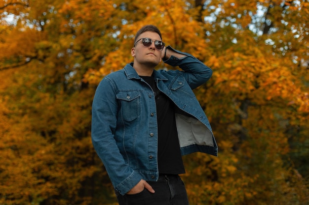 Stylish handsome young hipster guy with black sunglasses and a blue denim shirt walking in the countryside in an autumn forest with bright yellow foliage