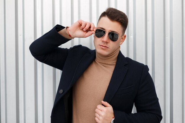 Stylish handsome young hipster man in a fashionable jacket in a knitted vintage sweater in trendy sunglasses with a stylish hairstyle poses outdoors near a metal wall