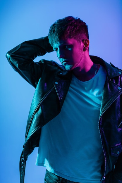 Stylish handsome young man with haircut in trendy outerwear with white t-shirt and fashion vintage black leather jacket against blue and pink light in studio. Multi-colored portrait of guy