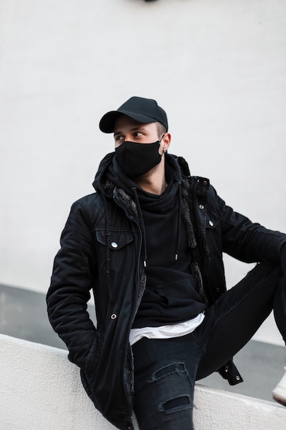 Stylish handsome young man with a medical mask in a fashionable cap with a black jacket and a hoodie sits on the street near a gray wall