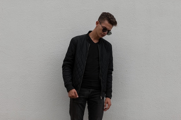 Stylish handsome young man with sunglasses in black fashionable clothes with a jacket near a gray wall on the street
