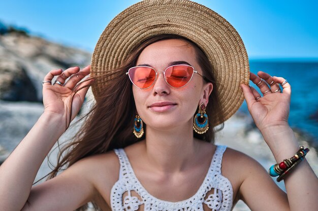 Stylish happy beautiful brunette woman with closed eyes wearing a straw hat, big earrings and red glasses enjoying summertime