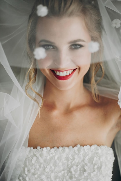 Photo stylish happy bride posing under veil and smiling in soft light near window in hotel room gorgeous sensual bride portrait morning preparation before wedding ceremony