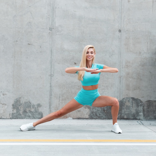 Stylish happy fitness model woman with wireless earphone in fashion blue sportswear with white shoes doing stretching and work out on the street near concrete wall