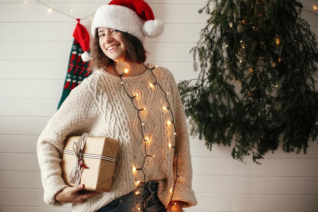 写真 現代のクリスマス ツリー ライトとストッキングの背景にクリスマス ギフト ボックスを保持しているサンタ帽子と居心地の良いセーターでスタイリッシュな幸せな女の子プレゼントを保持し、笑顔の若い流行に敏感な女性