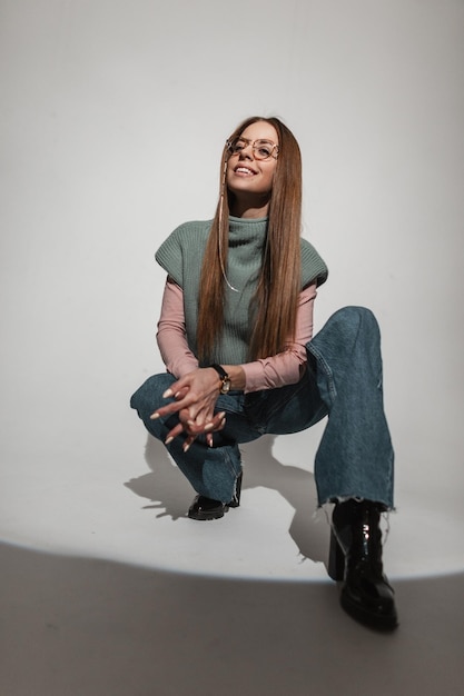 Stylish happy woman model in vintage fashion outfit with knitted top fashionable wide bell bottom jeans poses and sits in studio