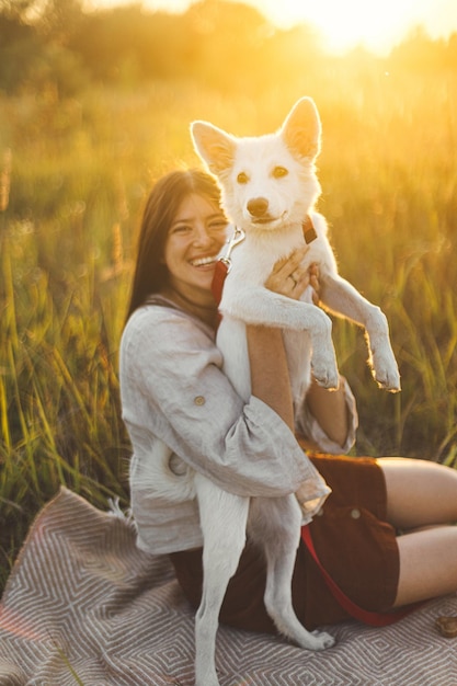 Stylish happy woman playing with her white dog on blanket in warm sunset light in summer meadow