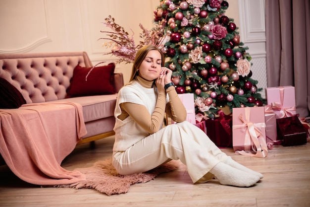A stylish happy young woman in a fashionable white knitted suit is sitting on a soft rug in a cozy room near a Christmas tree Fashionable girl joyful model rests in the Christmas studio