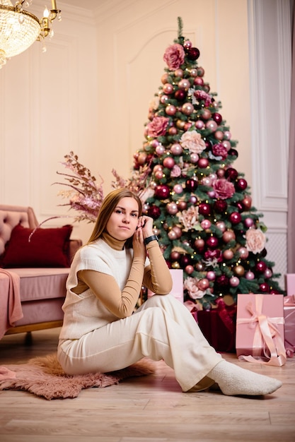 A stylish happy young woman in a fashionable white knitted suit is sitting on a soft rug in a cozy room near a Christmas tree Fashionable girl joyful model rests in the Christmas studio