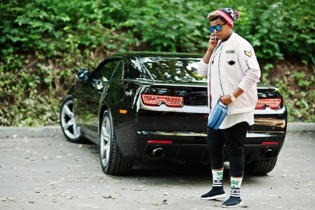 Stylish hipster arab man guy in hat and sunglasses posed\
outdoor in street against his black muscle car and speaking on\
mobile phone