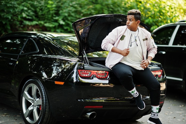 Stylish hipster arab man guy posed outdoor in street and sitting on the trunk his black muscle car Rich black man