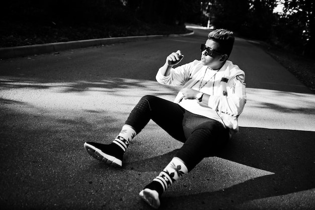 Stylish hipster arab man guy in sunglasses posed outdoor in street at sunset sitting on road in shadows