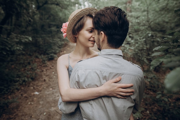 Stylish hipster bride and groom walking and dancing in green summer forest happy couple in love modern outfit relaxing at park girl in dress and straw hat with peony rustic wedding moment