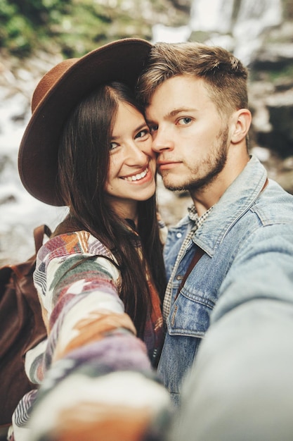 スタイリッシュな流行に敏感なカップルは、selfie を取って山の森の滝で笑顔ハンサムな男と新婚旅行で抱きしめる帽子で幸せな女は一緒に夏休みを旅行します。