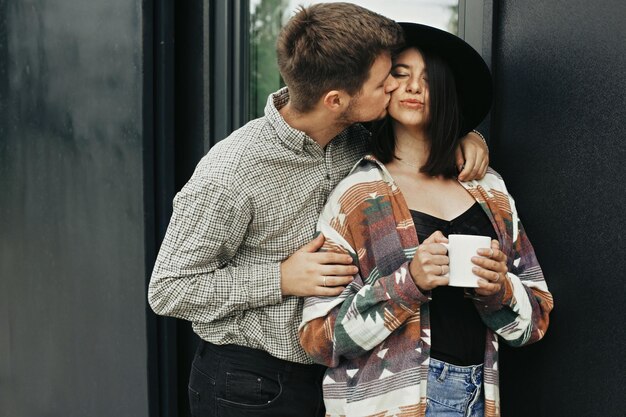 Stylish hipster couple with morning coffee relaxing on\
background of modern cabin and big windows in mountains happy young\
family enjoying morning at new home in woods travel and\
vacation