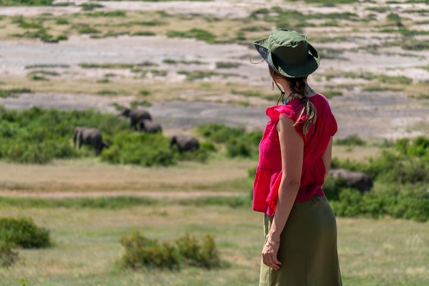 모자를 입은 세련된 스터 소녀가 사바나에서 코끼리의 배경으로 고 은 티셔츠를 입은 행복한 젊은 여성이 은 산을 탐험합니다.