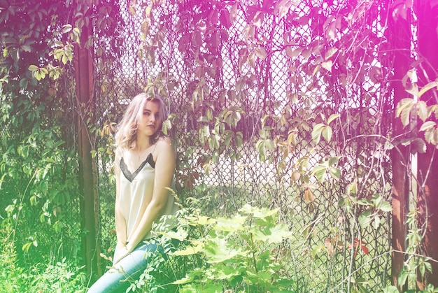 Stylish hipster girl is standing near the ivy leaves wall outdoors Fashionable photoshoot for young women Blonde short hair and cool glasses