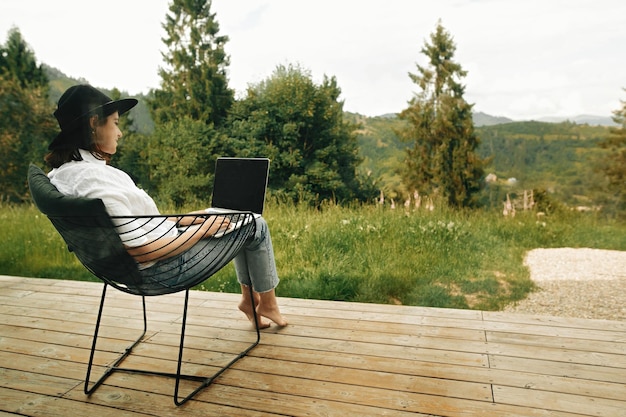 Photo stylish hipster girl sitting with laptop on terrace with view on woods young happy woman in hat using laptop shopping or working online from home outdoors freelance and freelancer