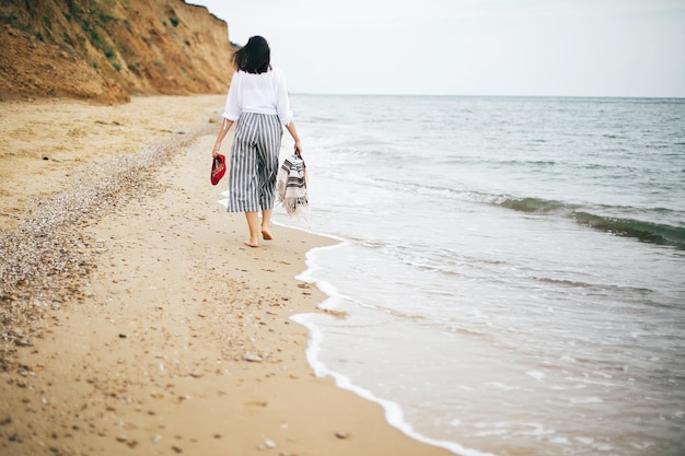 Foto elegante ragazza hipster che cammina a piedi nudi sulla spiaggia tenendo borsa e scarpe in mano felice donna boho che si rilassa in mare godendosi una passeggiata sull'isola tropicale vacanze estive spazio per il testo
