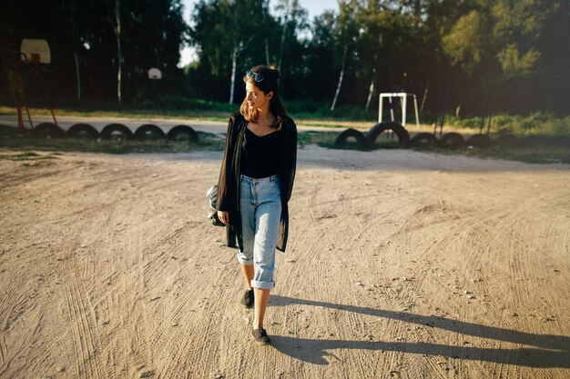 Stylish hipster girl walking in sunny street atmospheric moment Fashionable cool woman with black sunglasses and denim jeans relaxing at playground Selective focus Retro effect