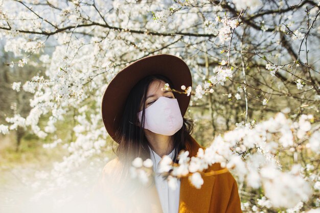 Стильная девушка-хипстер в маске для лица и наслаждается цветущими вишневыми деревьями в солнечном весеннем парке Молодая красивая женщина устала от карантина дома и отдыхает на улице в маске ручной работы
