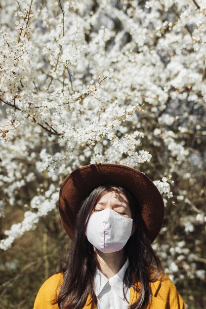 Stylish hipster girl wearing face mask and enjoying blooming cherry trees in sunny spring park alone Young beautiful woman tired of quarantine in home and relaxing outside in handmade mask