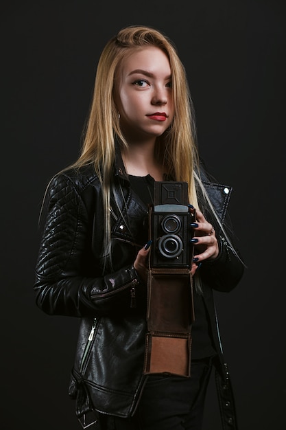 Stylish hipster girl wears black leather jacket and holding old vintage photo camera on black background