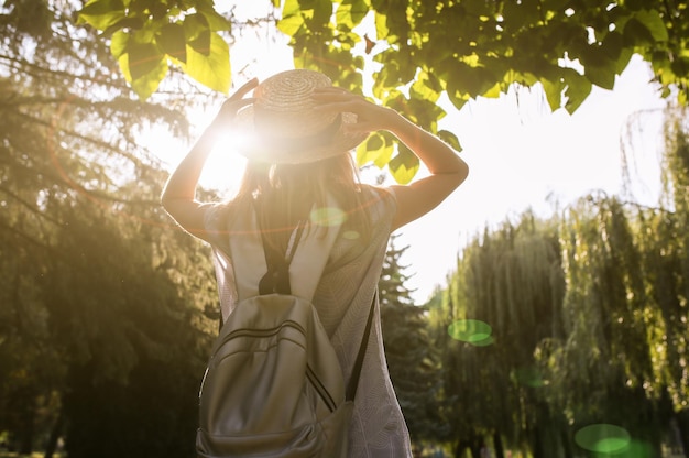 Foto stylish hipster ragazza con un cappello di paglia è in piedi all'aperto donna in abito blu zaino grigio argento concetto di vacanza di viaggio foto stile di vita estivo sfumature di sole e alberi