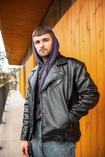 Stylish hipster street guy in a fashion black leather jacket and hoodie standing next to a wooden yellow wall