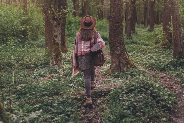 夕方の日差しの中で森の中を歩くバックパックを持った帽子をかぶったスタイリッシュな流行に敏感な旅行者の女の子