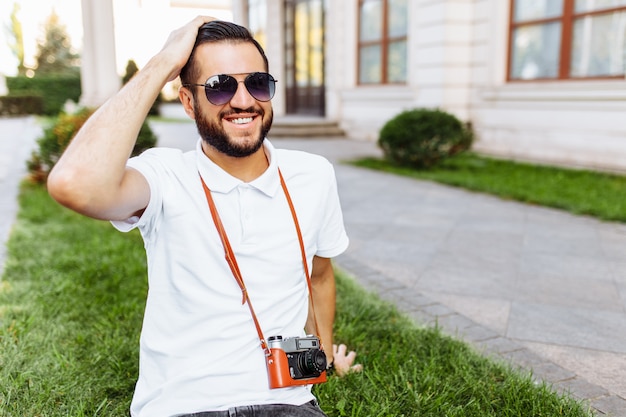 Elegante hipster con una macchina fotografica al collo, seduto sul prato nel parco