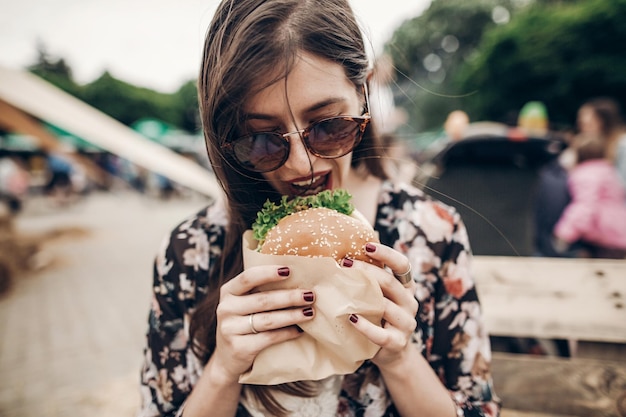 Stylish hipster woman holding juicy burger and eating boho girl with hamburger smiling at street food festival summertime summer vacation travel space for text