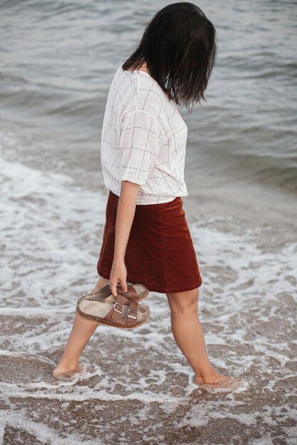 Photo stylish hipster woman walking barefoot on sandy beach with sea waves in the evening calm moment