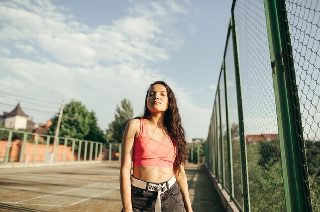 Stylish Hispanic girl stands on a playground near a fence off the grid