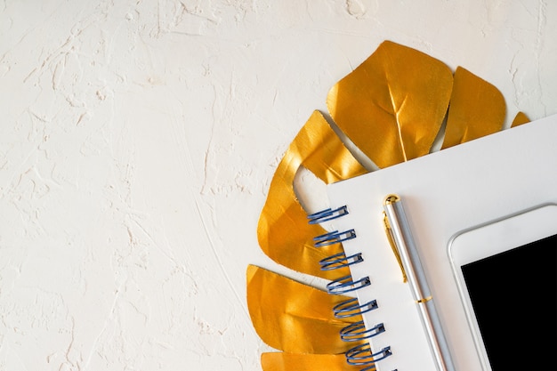 Stylish home office desk with  paper notepad, white smartphone, golden pen and tropical monstera leaf. Flat lay, top view.