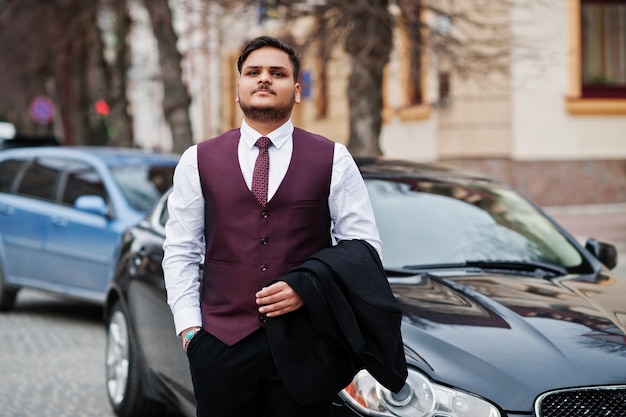 Stylish indian businessman in formal wear vest suit standing\
against black business car on street of city